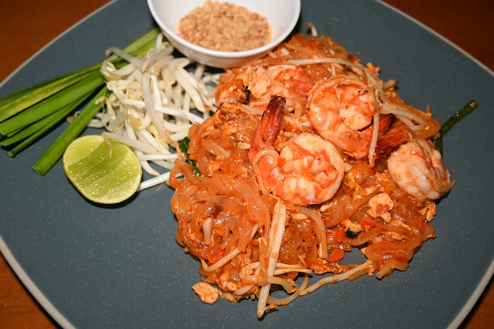 Symbolbild: Pad Thai mit Garnelen, Limette, Sojasprossen und Erdnüssen auf dunklem Teller